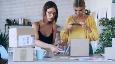 İki güzel serbest çalışan kadının akıllı telefonlarıyla fotoğraf çekerken yeni başladıkları küçük işletmelerde karton kutuları paketleyip mühürledikleri bir video.. 