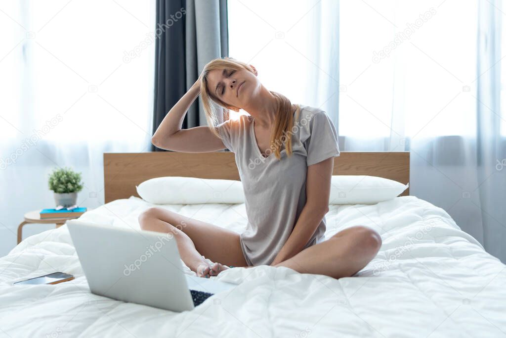 Shot of beautiful young woman with neck pain working with laptop sitting on the bed at home.