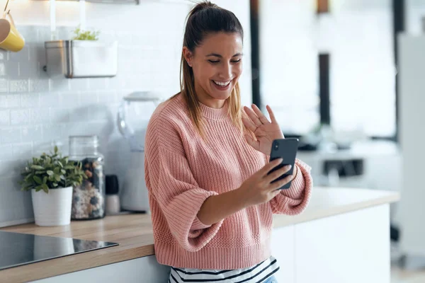 自宅で台所でスマートフォンでビデオ通話をしながら手を振ってかなり若い女性のショット — ストック写真
