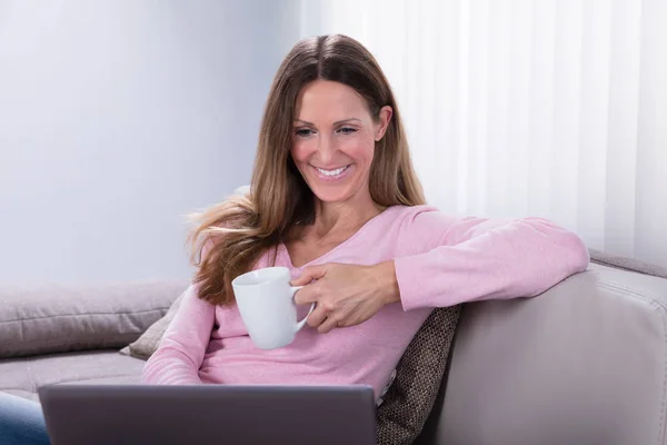 微笑成熟的女人坐在沙发上拿着咖啡杯看着笔记本电脑 — 图库照片