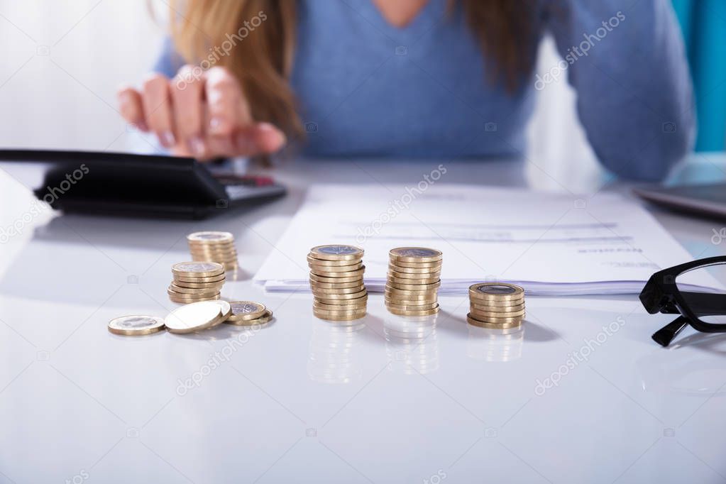 Close-up Of Businesswoman Calculating Invoice With Stacked Of Golden Coins On White Desk In Office