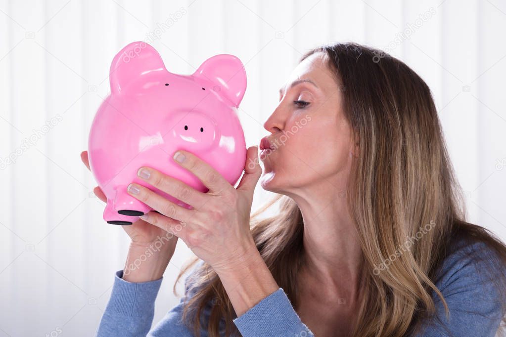 Side View Of Mature Woman Kissing Piggybank Against Blinds