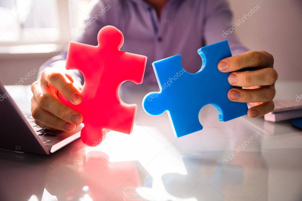 Close-up Of Businessperson's Hand Holding Two White Jigsaw Puzzle Over Desk