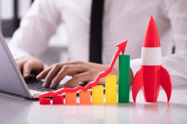Rocket And Multi Colored Graph With Arrow In Front Of Businessperson Working On Laptop clipart