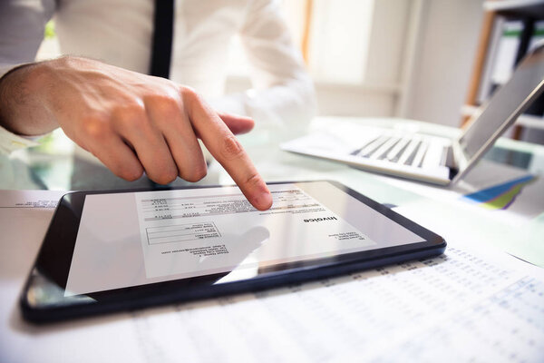 Close-up Of A Businessperson's Hand Analyzing Bill On Digital Tablet Over Desk