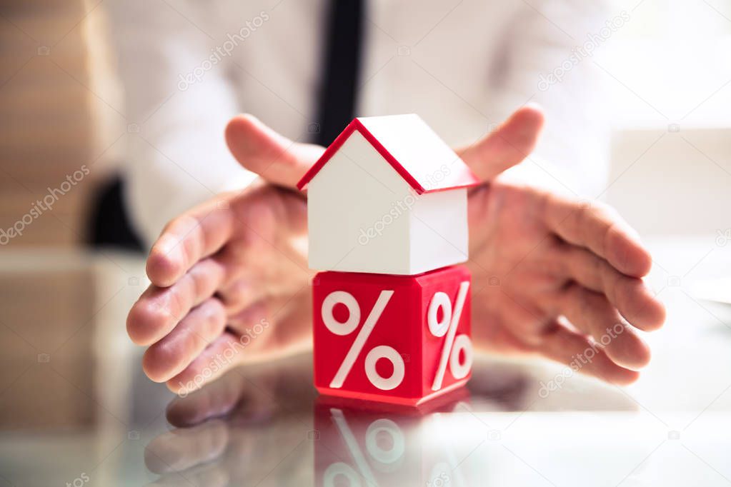 Close-up Of A Person's Hand Protecting House Mode Over Red Cubic Clock With Percentage Symbol