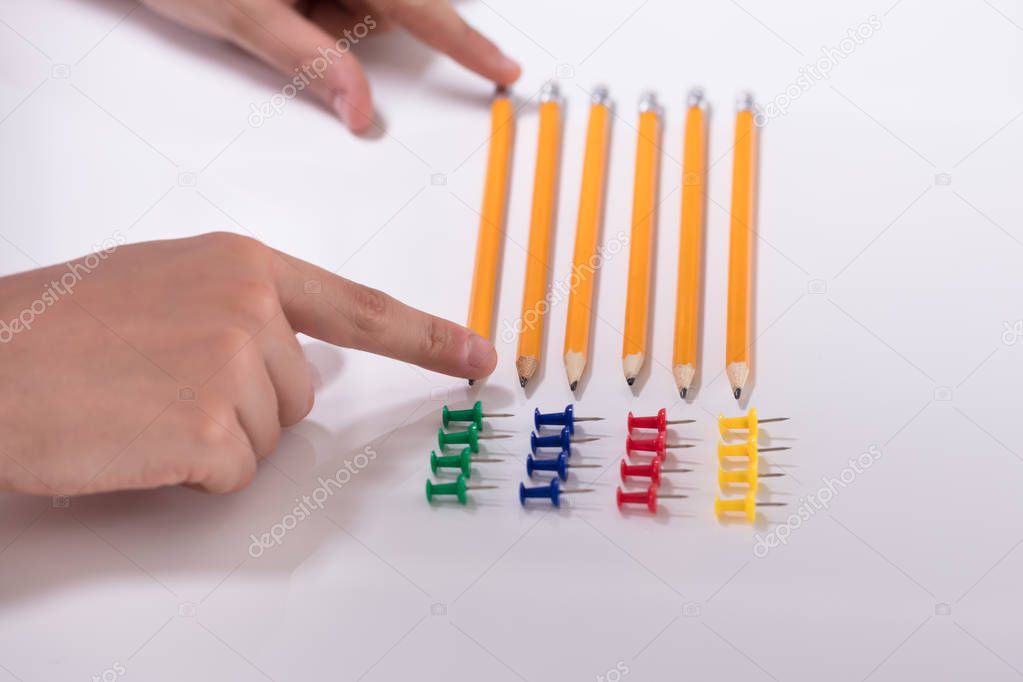 A Person's Hand Arranging Pencils And Multi Colored Pushpins In A Row On White Background