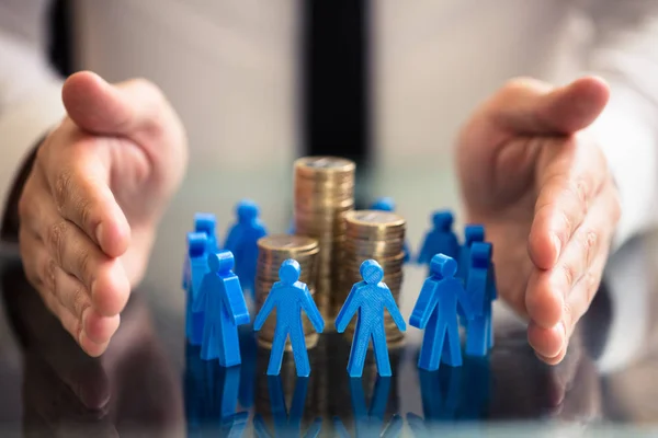 Businessperson Hand Protecting Blue Human Figures Surrounding Stacked Golden Coins — Stock Photo, Image
