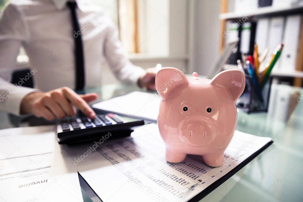 Close-up Of A Piggybank In Front Of Businessperson's Hand Using Calculator At Workplace