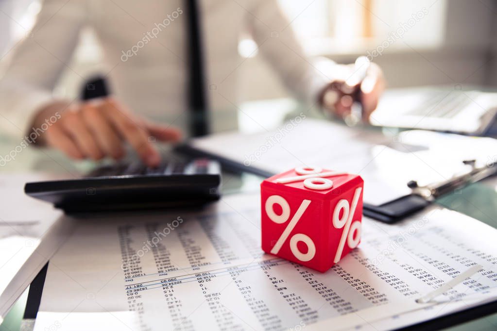 Close-up Of Red Cubic Block With Percentage Symbol In Front Of Businessperson Calculating Invoice