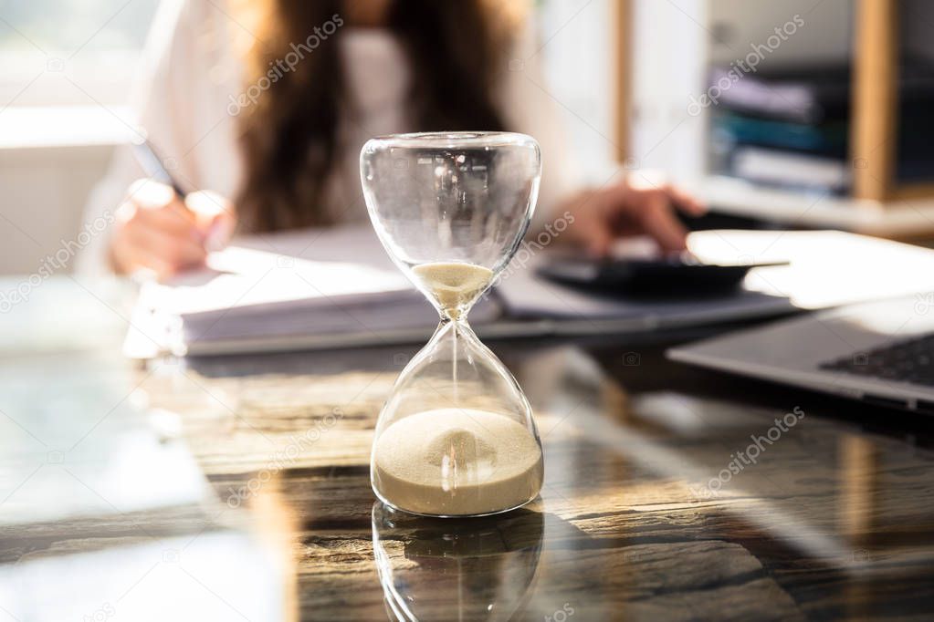 Close-up Of A Hourglass In Front Of Businesswoman Working In Office