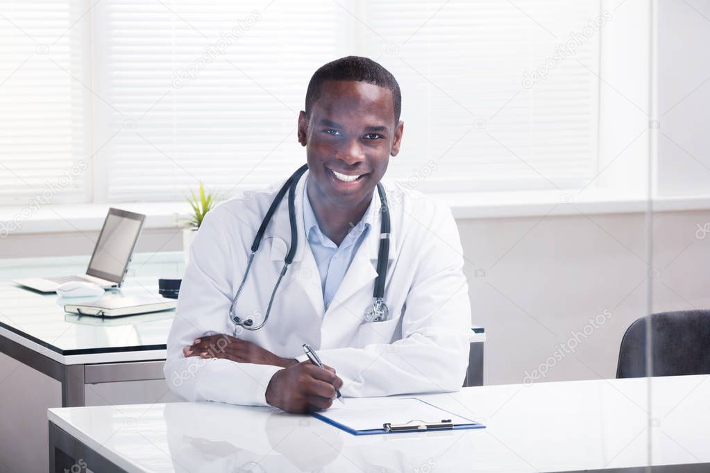 Close-up Of A Smiling Male Doctor With Clipboard And Pen Sitting In Clinic