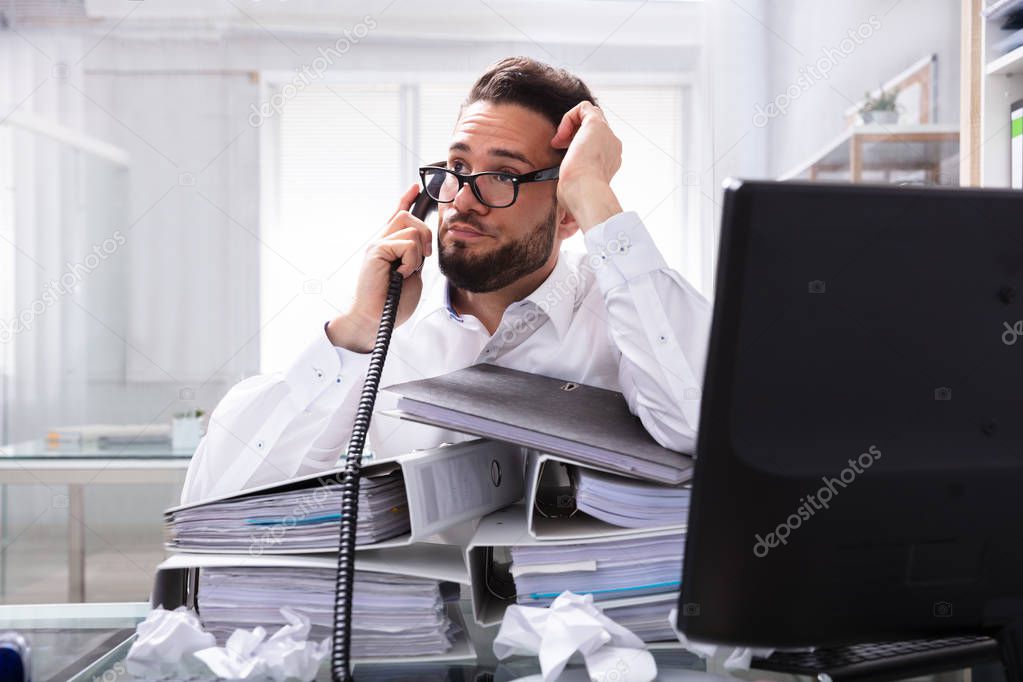 Overworked Businessman Talking On Landline With Stack Of Folders On Desk