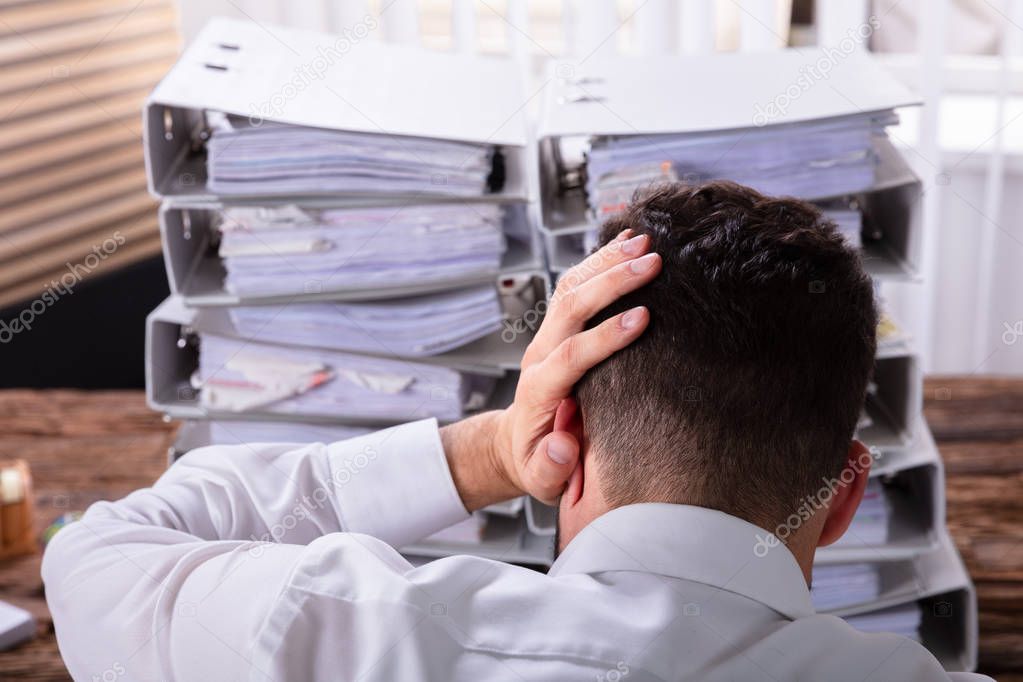 Close-up Of Stressed Businessman With Hand On Head In Front Of Stacked Folders