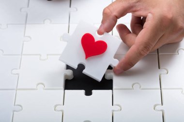 Person's hand placing last piece with red heart into white jigsaw puzzle