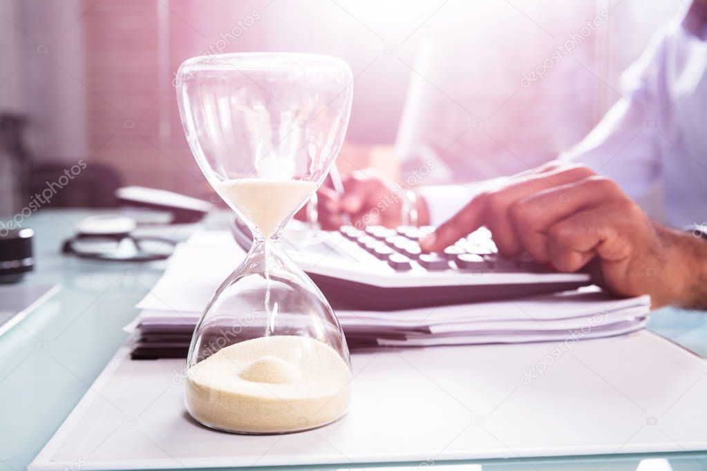 Close-up Of Hourglass In Front Of Businessperson's Hand Calculating Invoice Using Calculator