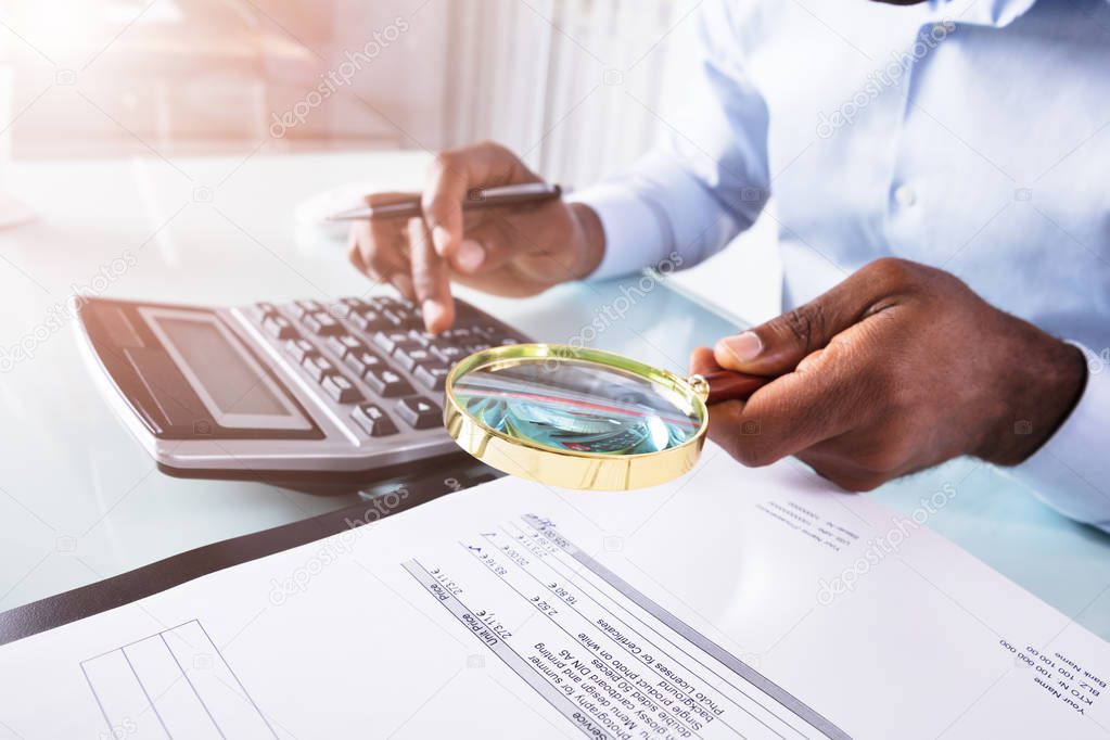 Close-up Of A Businessman Holding Magnifying Glass Over Invoice Using Calculator
