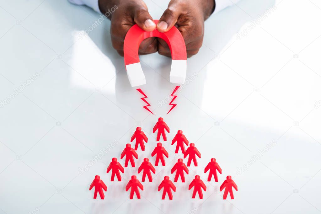 Close-up Of A Human Hand Attracting Red Human Figures With Horseshoe Magnet On White Background