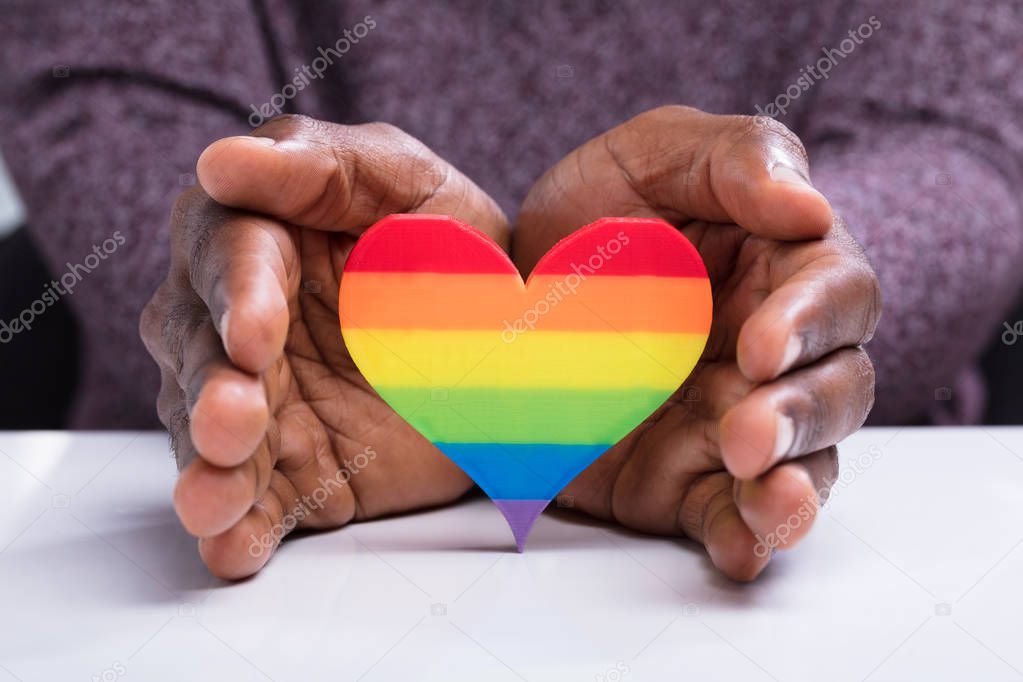 Close-up Of A Man's Hand Protecting Colorful Strip Heart Over The White Desk