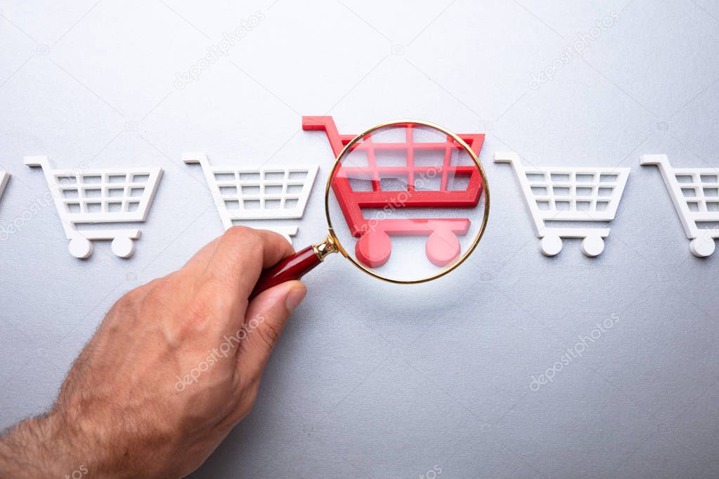 A Person Looking At Shopping Cart Through Magnifying Glass On Grey Background