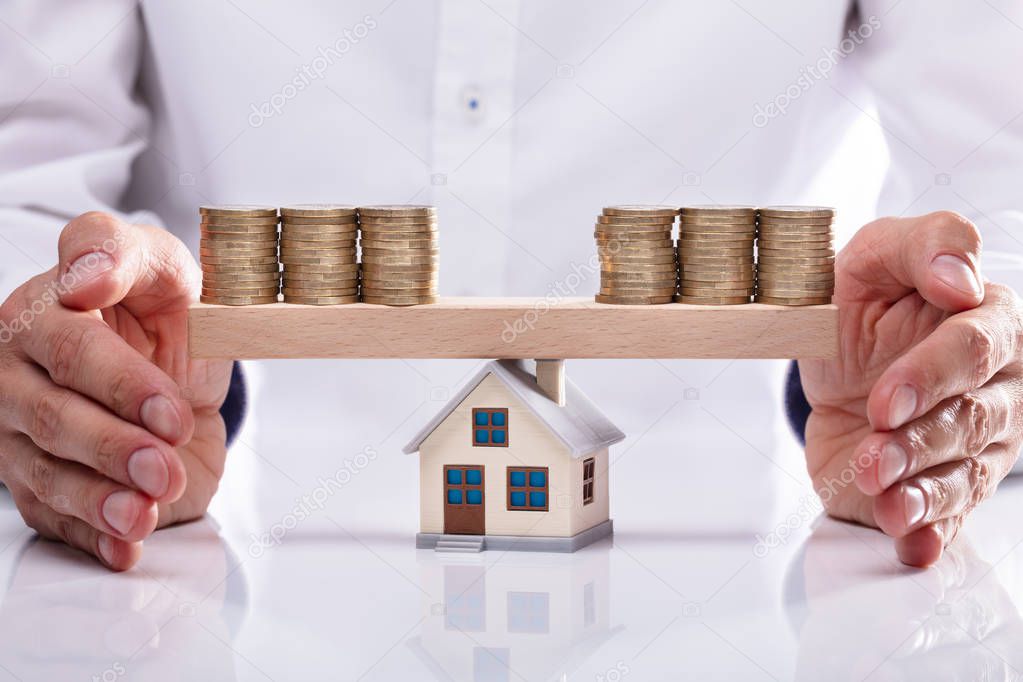 Businessman's Hand Protecting Stacked Coins On Seesaw With House Model