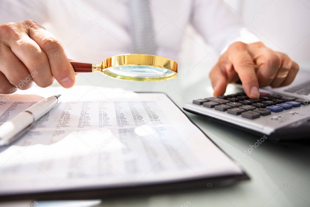 Businessman's Hand Using Calculator While Analyzing Financial Report With Magnifying Glass