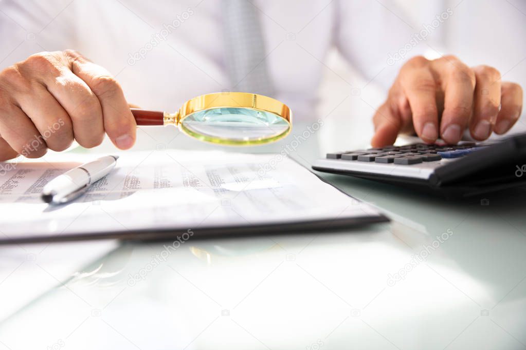 Businessman's Hand Using Calculator While Analyzing Financial Report With Magnifying Glass