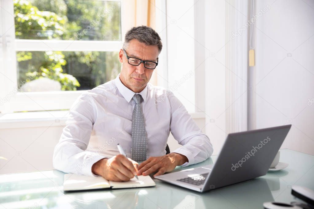 Mature Businessman Writing Schedule In Diary With Pen Over Desk