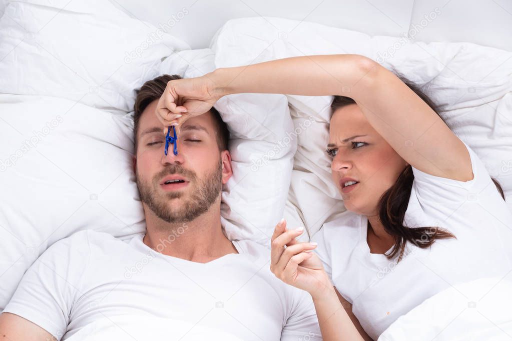 Frustrated Young Woman Trying To Stop Man's Snoring With Clothespin On Bed