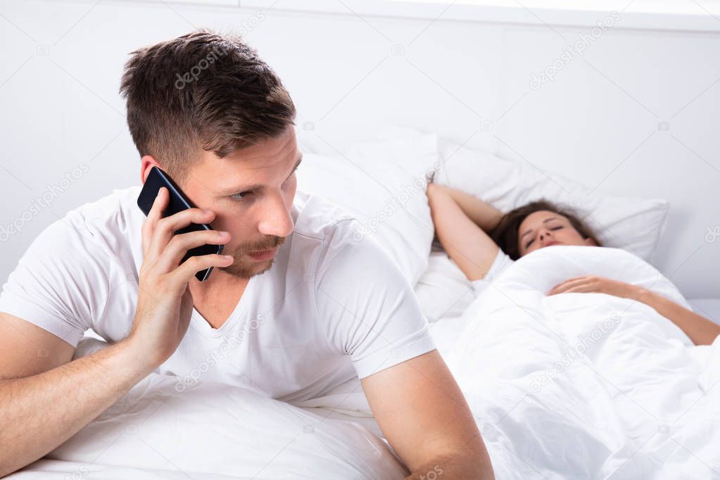 Young Man Talking On Mobile Phone While Her Wife Sleeping On Bed