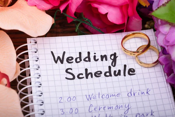 两个金色戒指与婚礼时间表在螺旋记事本周围的花朵 — 图库照片