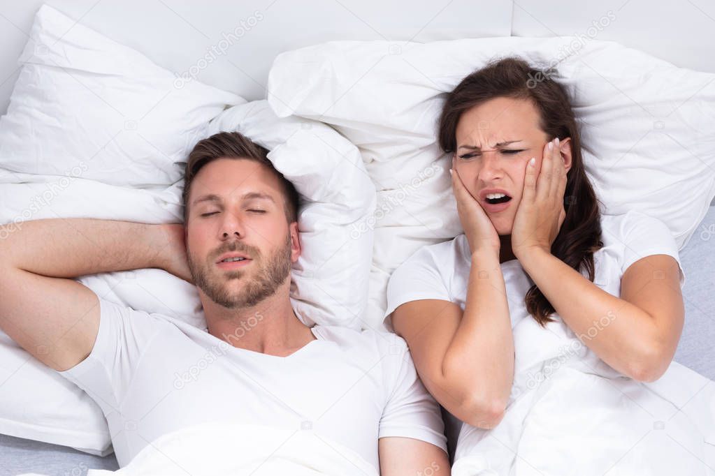 Disturbed Young Woman Covering Her Ears With Hands While Man Snoring On Bed