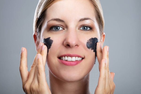 Portrait Of A Smiling Young Woman Applying Activated Charcoal Mask On Her Face