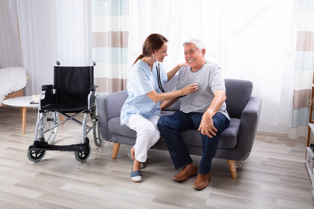 Female Nurse Checking Happy Senior Man Sitting On Sofa With Stethoscope