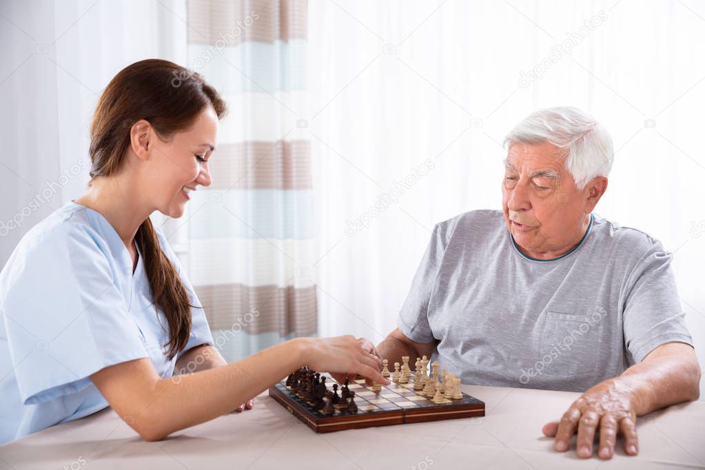 Young Female Caretaker Playing Chess With Senior Man At Home