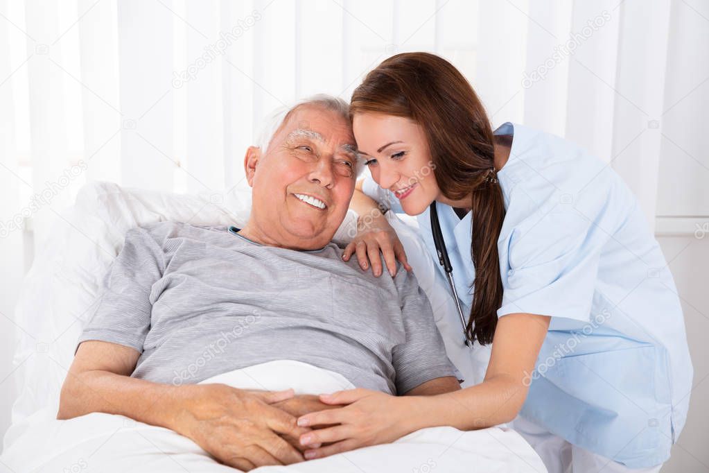 Nurse Looking At Happy Male Patient Lying On Bed In Clinic