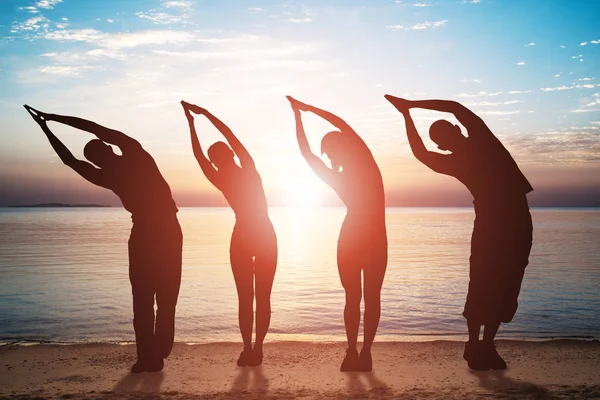 Silhouette Of People Doing Stretching Exercise On Sandy Beach At Sunset