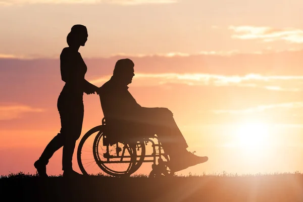 Silueta Mujer Ayudando Padre Discapacitado Silla Ruedas Parque Atardecer — Foto de Stock