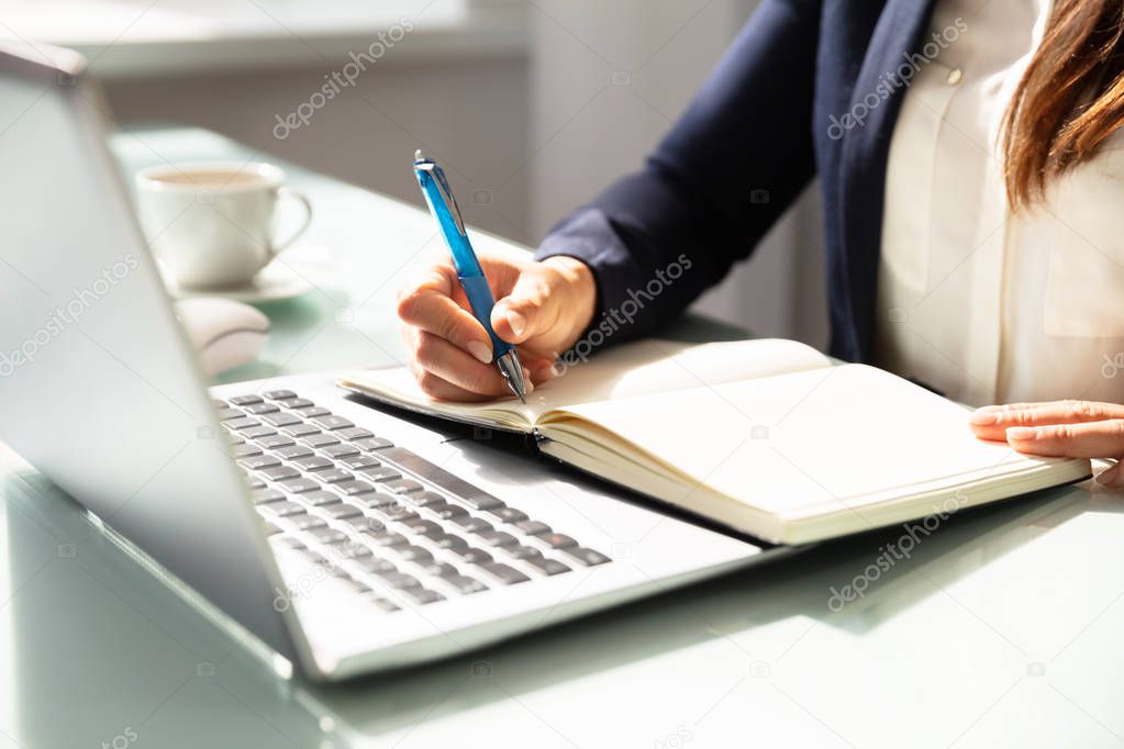 Close-up Of A Businesswoman's Hand Writing Note With Pen In Diary Over Desk