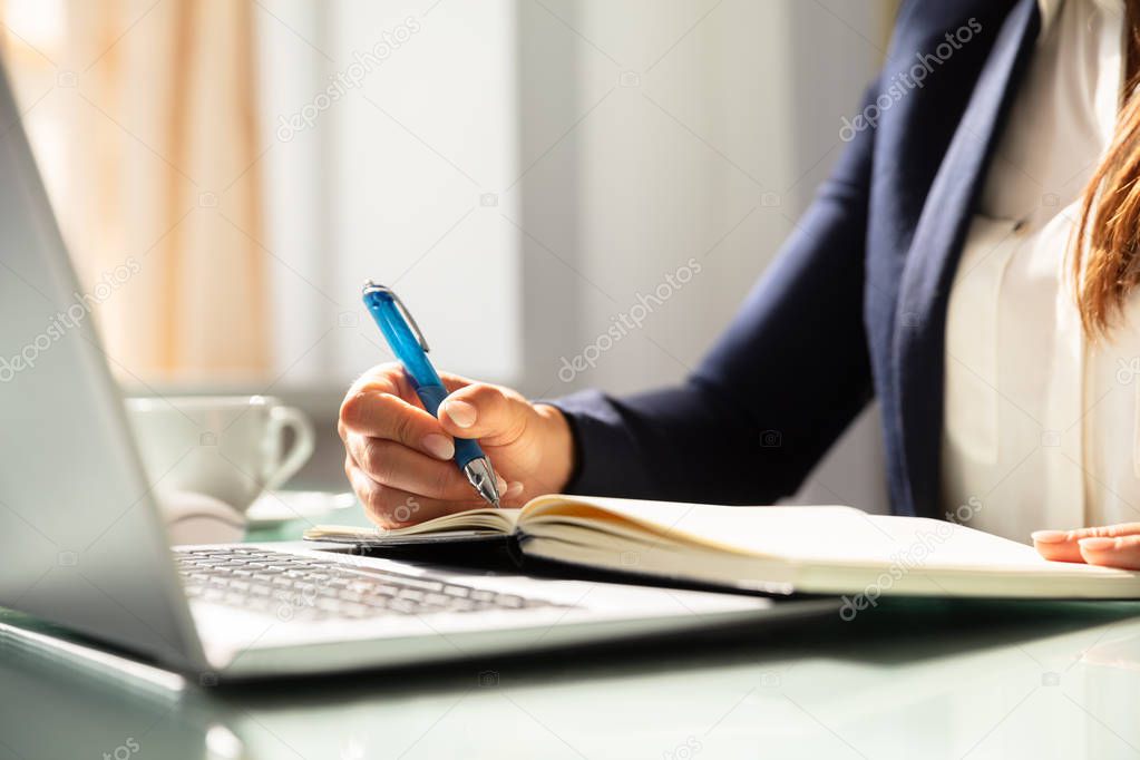 Close-up Of A Businesswoman's Hand Writing Note With Pen In Diary Over Desk