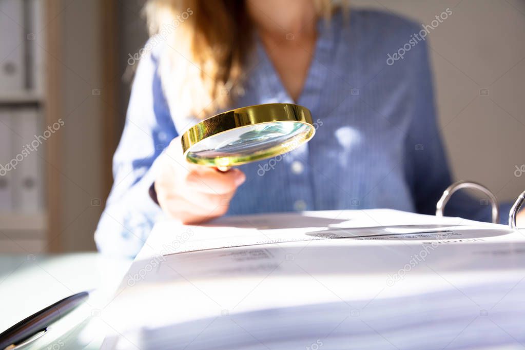 Close-up Of A Businesswoman's Hand Checking Invoice With Magnifying Glass Over Desk