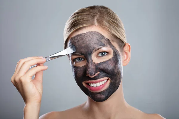 Junge Frau Trägt Aktivkohle Gesichtsmaske Mit Pinsel Auf Grauem Hintergrund — Stockfoto