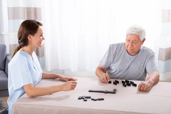Jonge Vrouwelijke Verzorger Zoek Naar Oudere Man Spelen Van Domino — Stockfoto
