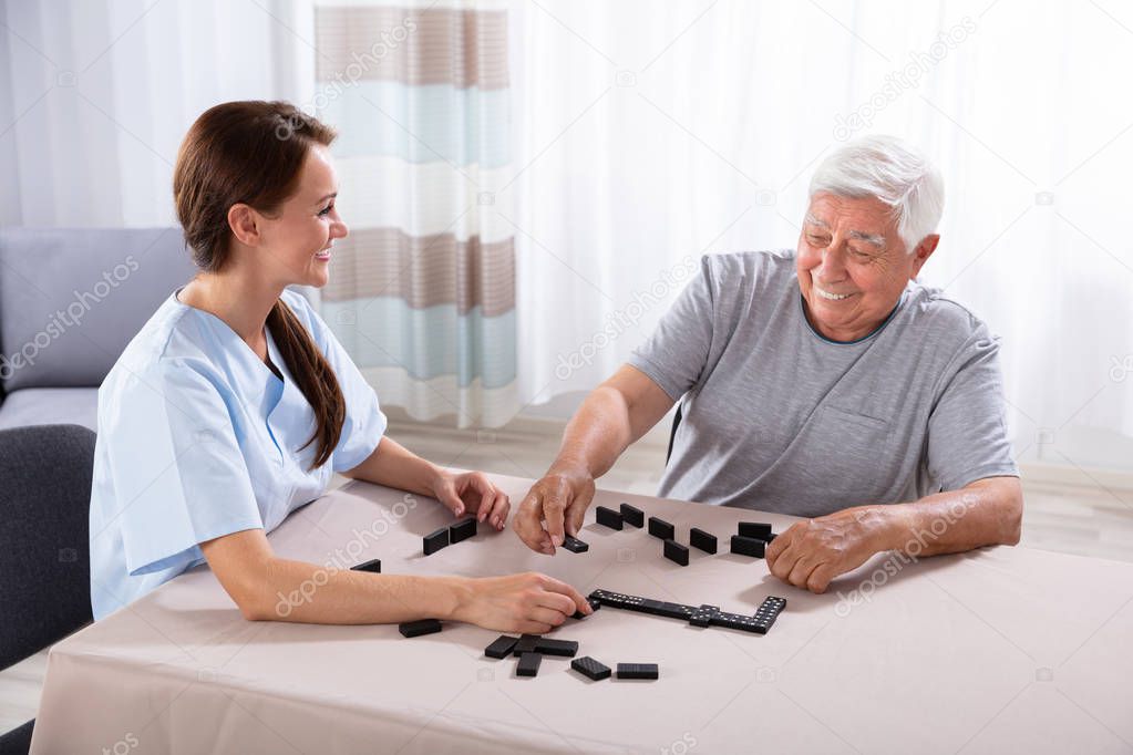 Young Female Caretaker Looking At Elder Man Playing Dominoes On Desk