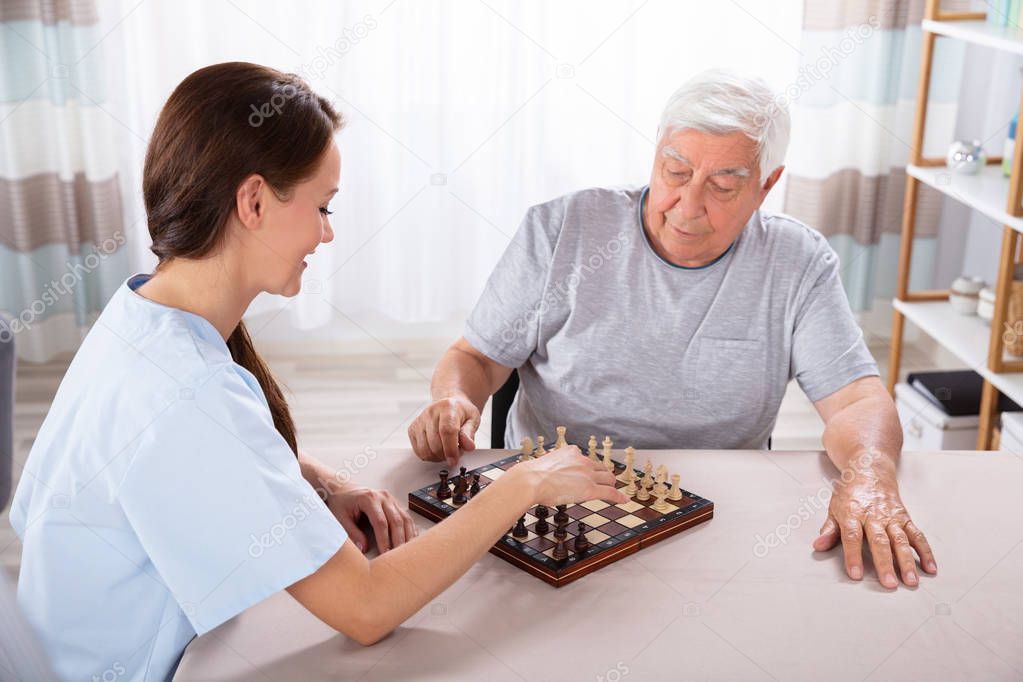 Young Female Caretaker Playing Chess With Senior Man At Home