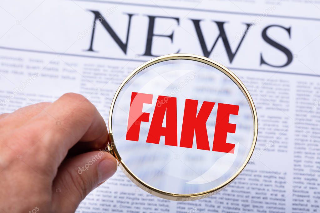 Man's Hand Examining Fake News On Newspaper Through Magnifying Glass