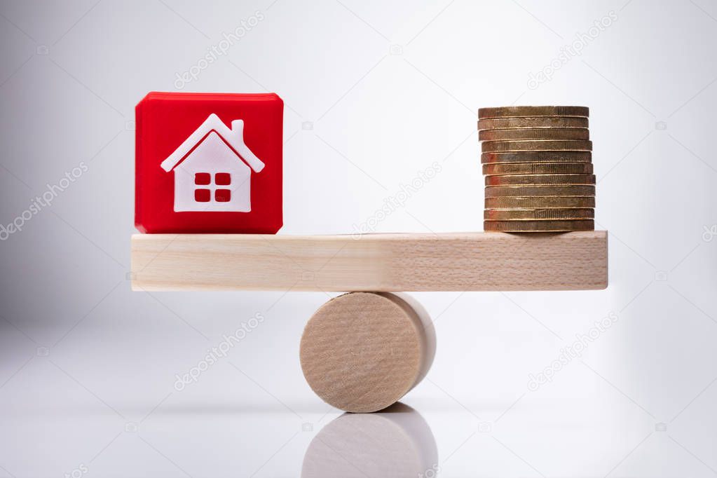 Red House Model Cubic Block And Stacked Golden Coins Balancing On Wooden Seesaw