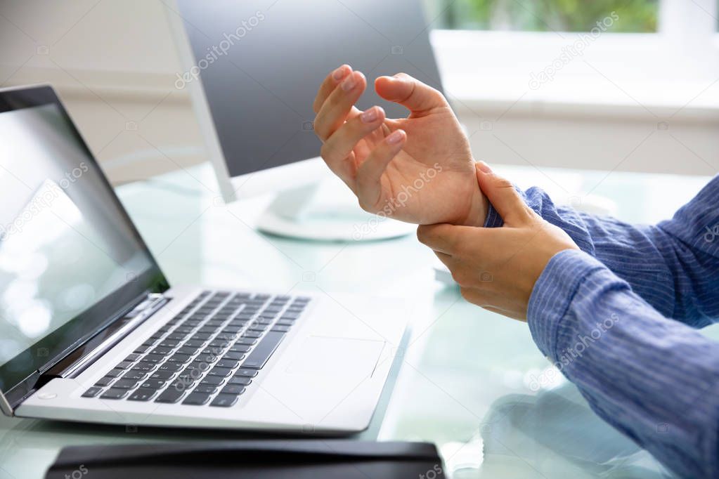 Close-up Of A Businesswoman's Hand Holding Her Painful Wrist With Laptop On Desk