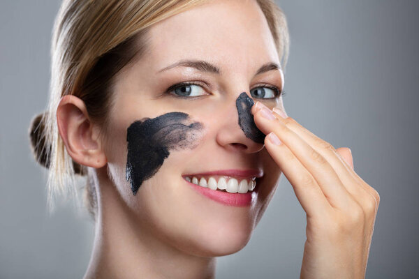 Portrait Of A Smiling Young Woman Applying Activated Charcoal Mask On Her Face