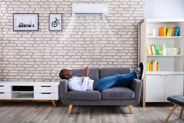 Young African Man Relaxing Sofa Operating Air Conditioner Remote Control — Stock Photo, Image